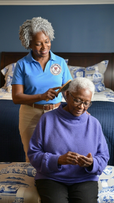 An older woman in a purple blouse sits in a chair while a female care giver stands behind her with a brush in her hand.