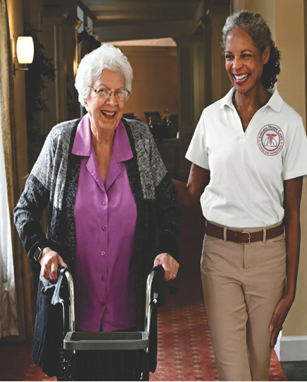 An older woman in a purple blouse walks behind a walker while a care giver walks next to her with a hand on the older woman's shoulder.