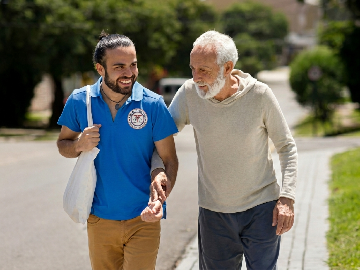 An older man and a male care giver walk on a sidewalk outdoors. Both are smiling. 