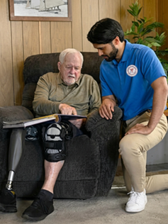 An older man sits in an arm chair looking at a book while a young male care giver kneels next to him. 